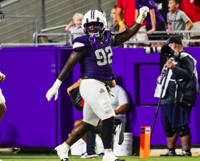 TCU defensive tackle Ansel Din-Mbuh, wearing a purple No. 92 football jersey, white pants and a white helmet, raises his right fist in celebration on the field during a game. Fans, photographers and purple stadium walls are visible in the background.