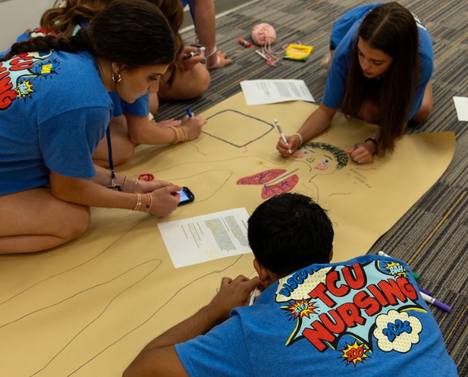 Three high school students are on the ground, surrounding a large paper with a drawing of a person. All students are wearing blue shirts that say 